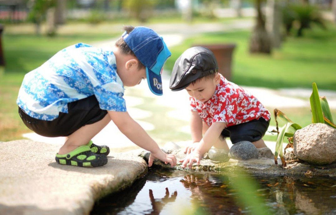 Two children outdoors studying something on the ground next to a pond.