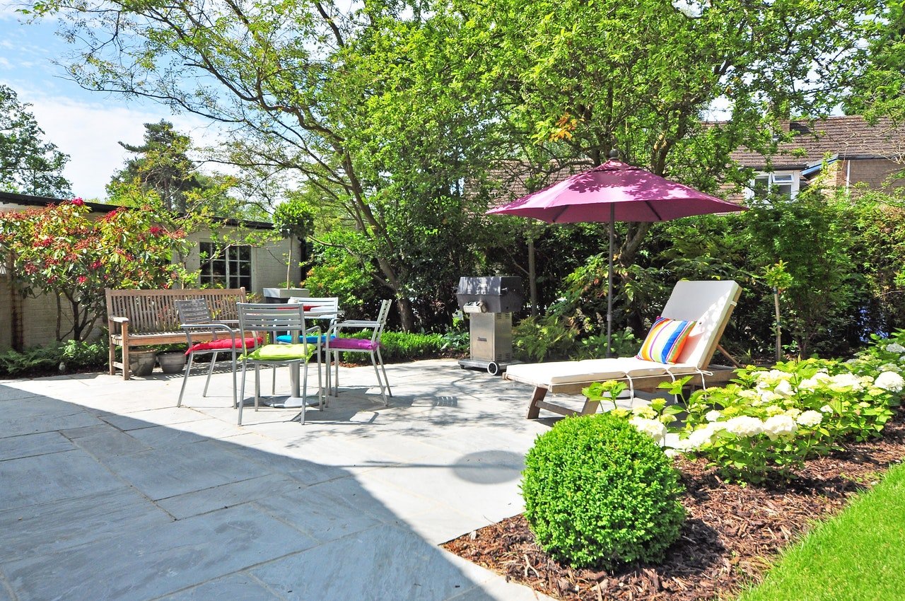 An image of a garden patio with various items of furniture, including sun lounger, table and chairs, and a bench. Plus parasol for shade.