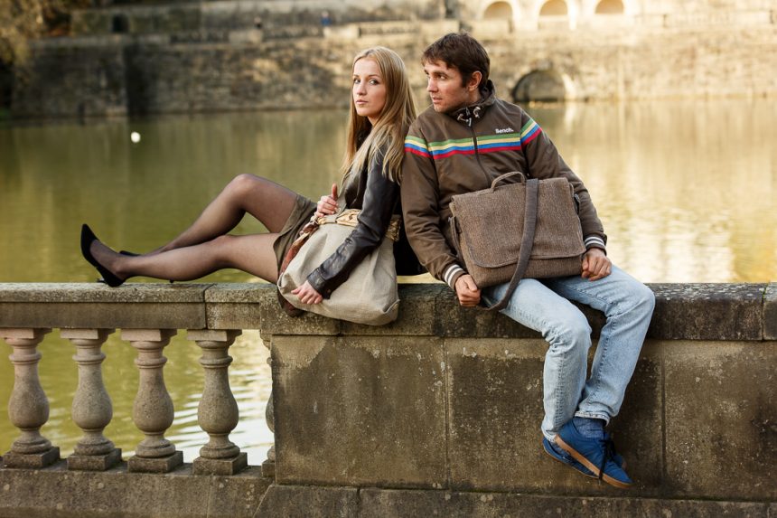 Messenger bag. Girl and boy sat on wall next to river. Both have a bag