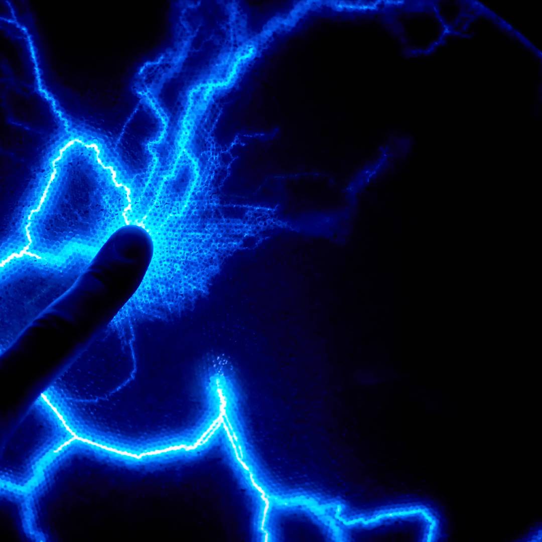 A silhouette of a finger touching a dark round object that’s putting of arcs of glowing blue static electricity.