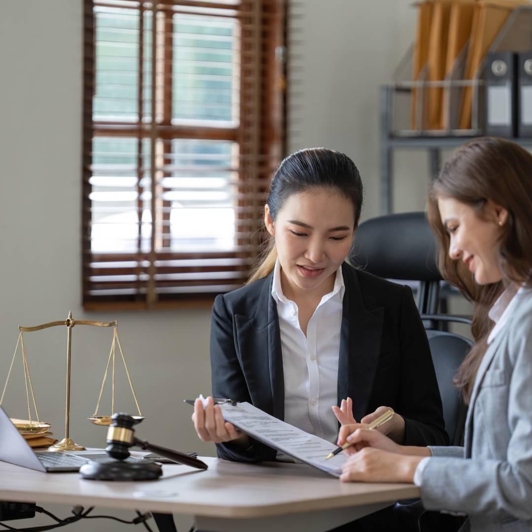 A lawyer sits at her desk with a client, reviewing a legal document on a clipboard. The client holds a pen.