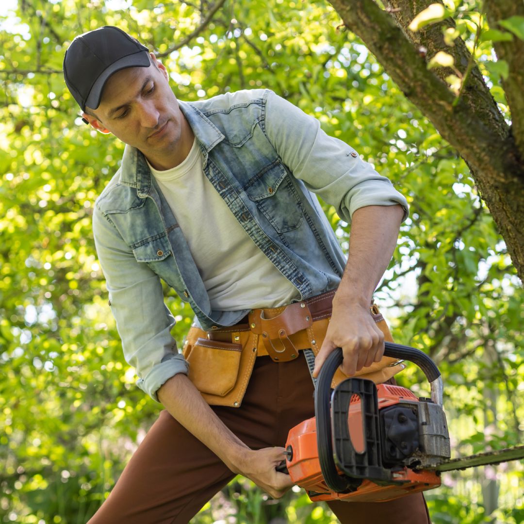 A man preparing to use a chainsaw to cut down a tree. He wears a baseball cap, a denim shirt, and a tool belt.