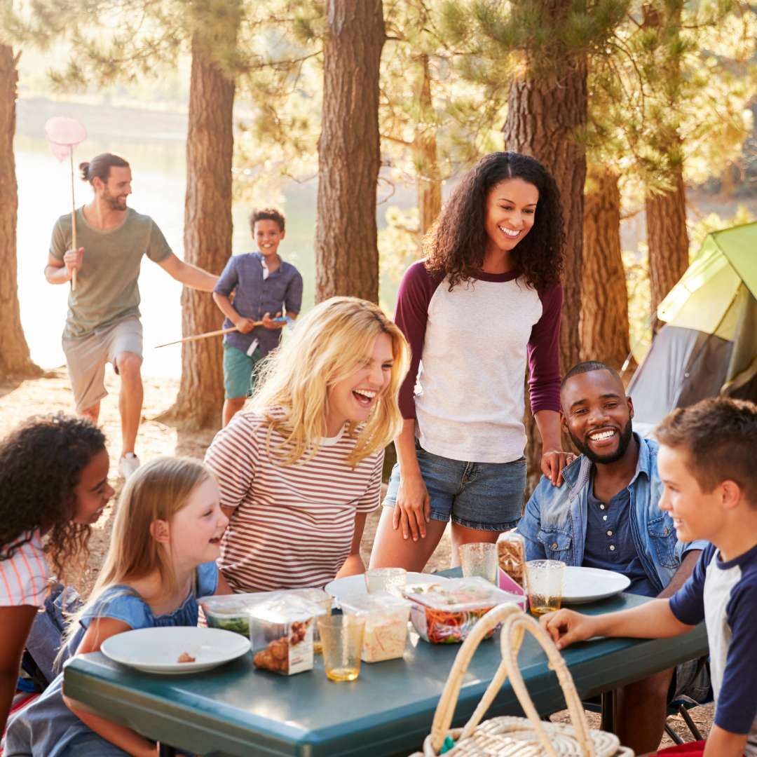 A group of several smiling people sitting around a picnic table while camping with a tent and trees in the background.