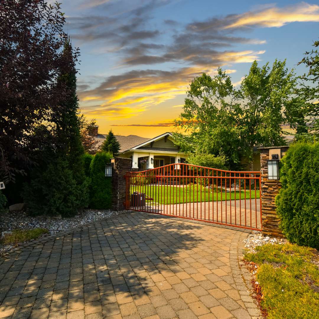 A brick driveway with a red metal gate guarding the entrance to a home. Decorative plants and gravel line the gate entrance.