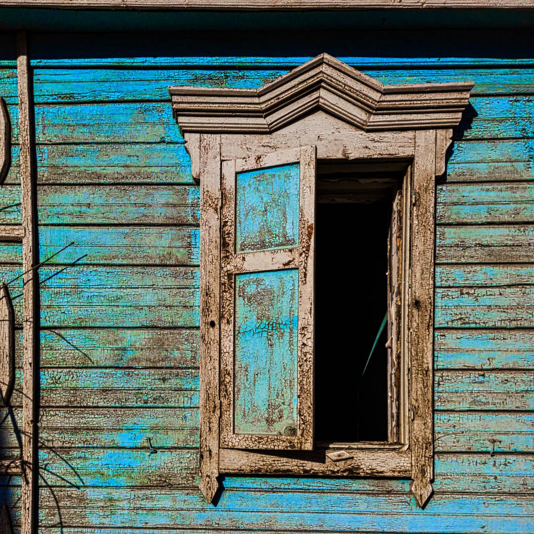 The side of a house has fading green paint. The wood's original color shows along with an old wooden window hanging open.