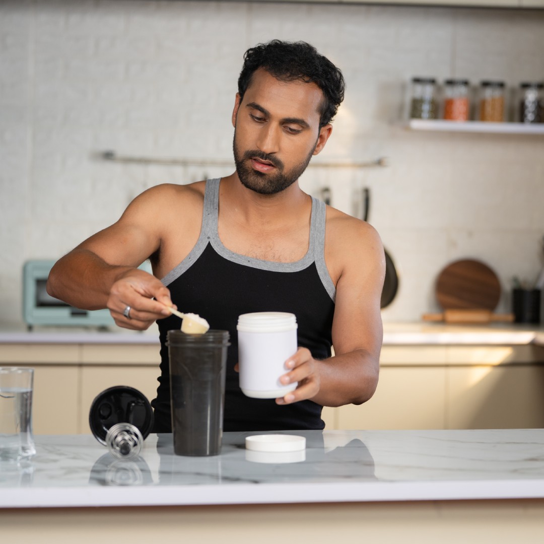 A man in a gray and black sleeveless shirt stands in his kitchen and adds powder to his drink with a measuring cup.