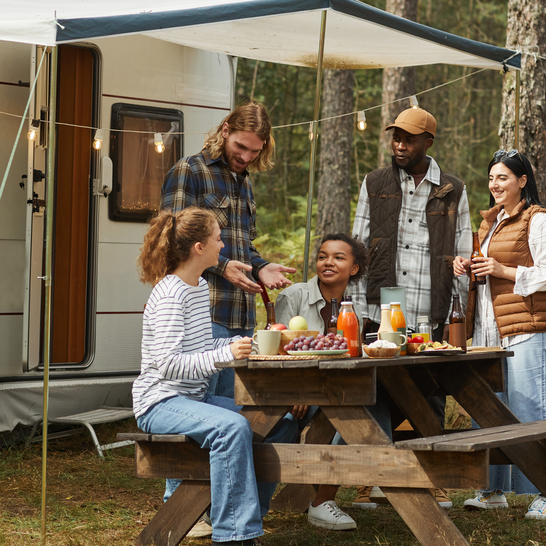 A group of people gathered around a picnic table at a wooded campsite. The table is filled with food and drinks.