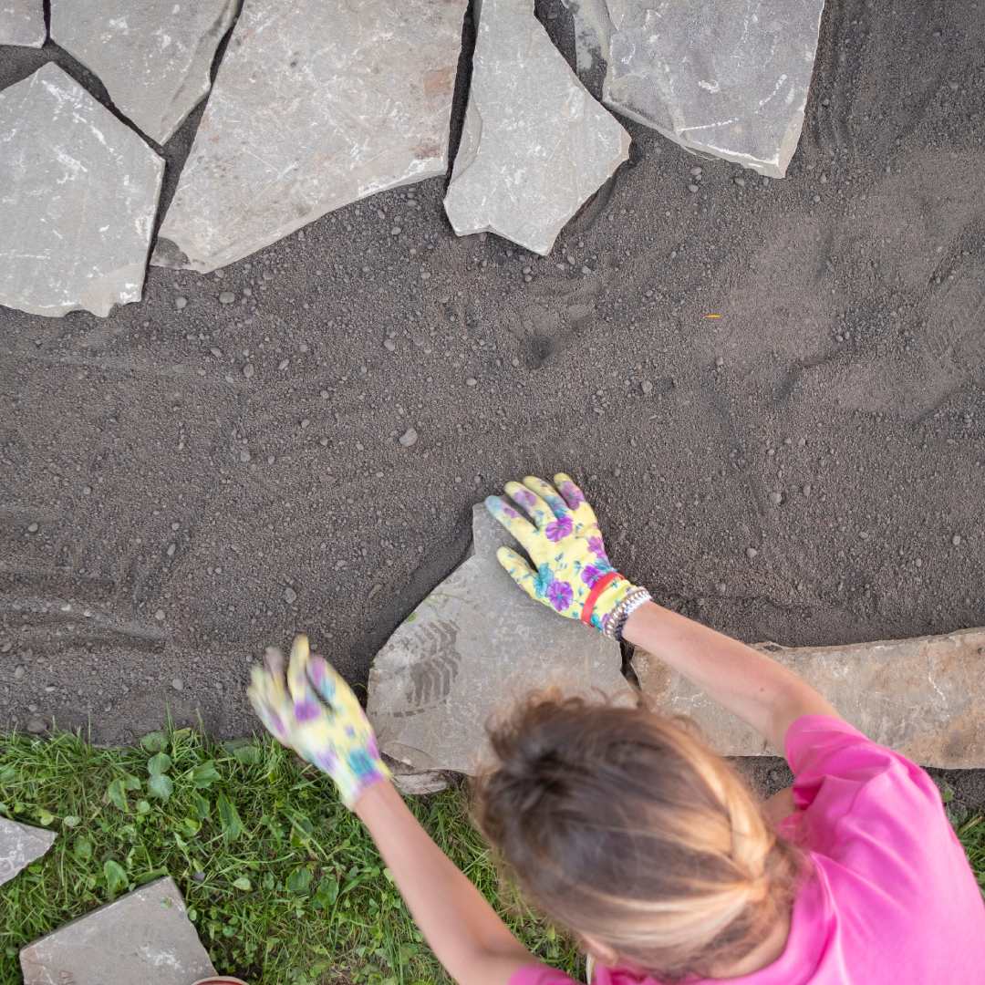 A woman wearing a pink shirt and colorful gloves laying large stones in her yard for a landscaping project.