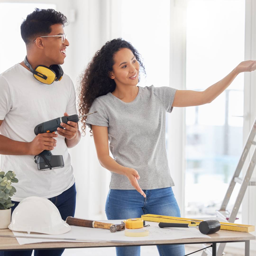 A couple stands in their home in front of a table that features a hard hat, tools, and plans. The husband holds a drill.