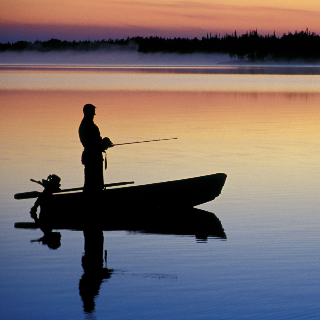 A man stands in his boat while fishing in a lake during early evening hours. A forest stands in the background.