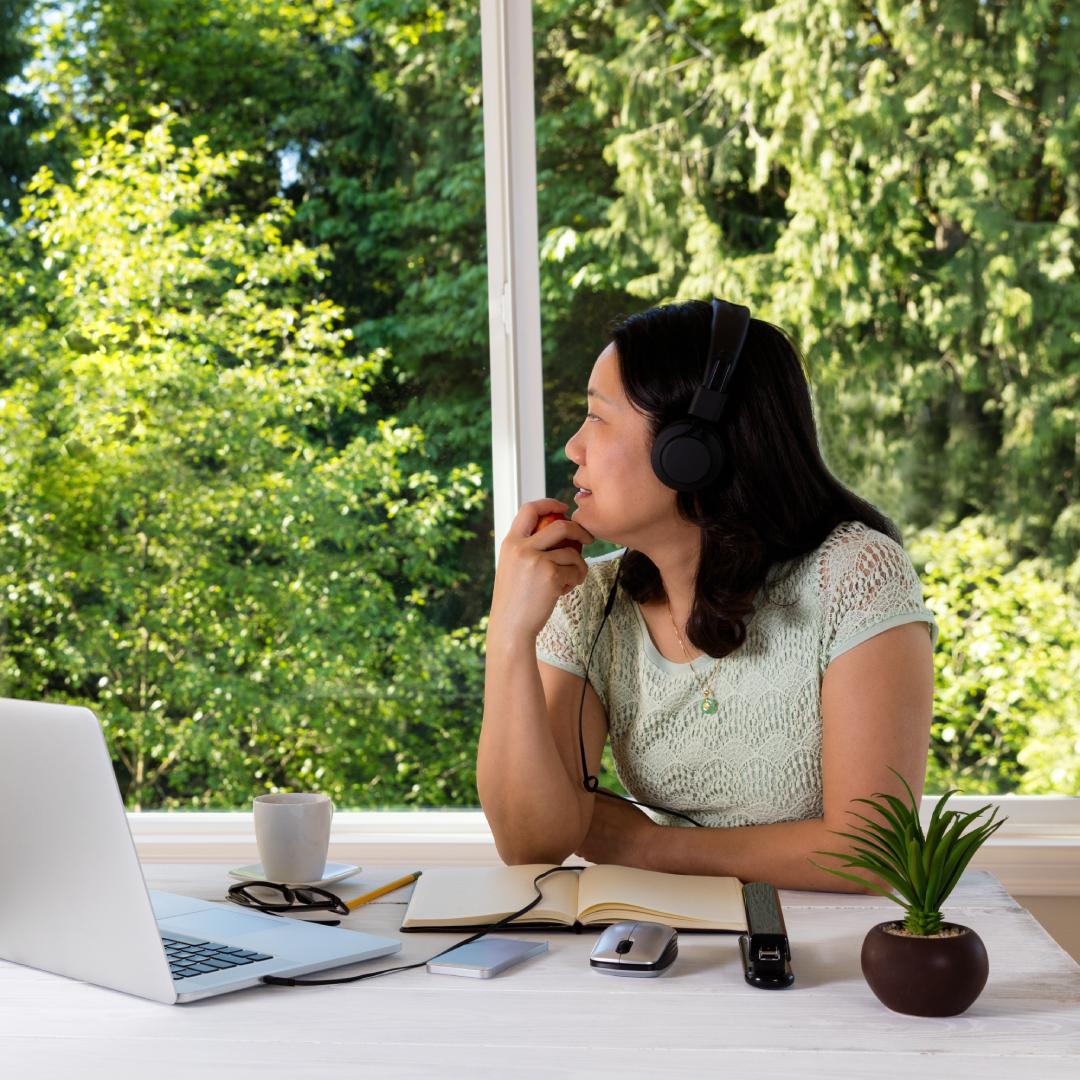 A woman wearing headphones sitting at her desk in her office at home. Her office has large windows with a view of trees.