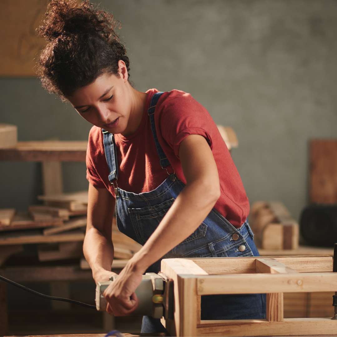 A young woman in denim overalls and a red tee shirt using an electrical belt sander to smooth a wooden stool.