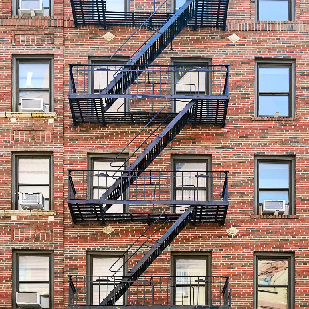 A brick residential apartment building shows five levels. Each level has a black metal fire escape platform and stairs.