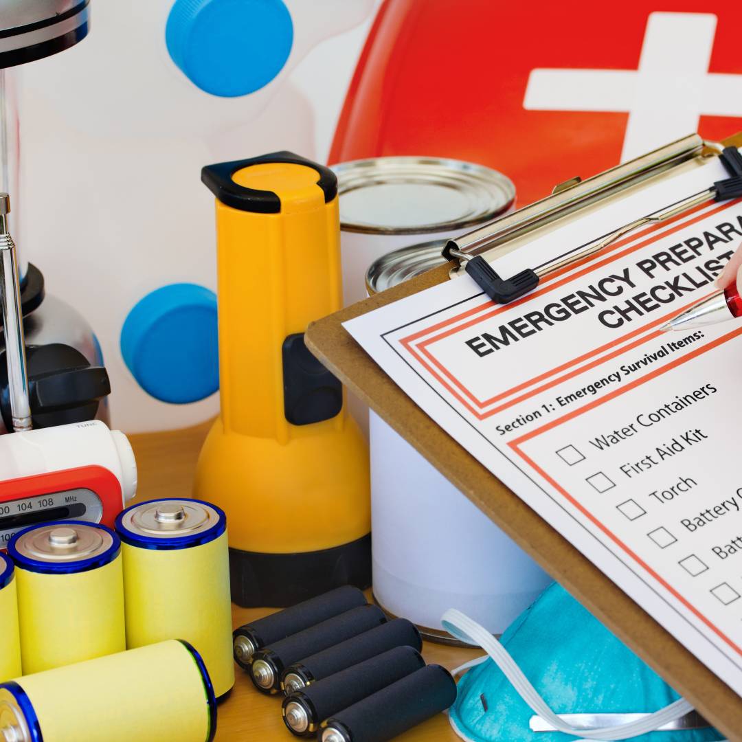 A person holds a clipboard with a paper that reads, "Emergency Preparation Checklist." Various supplies sit on a table.