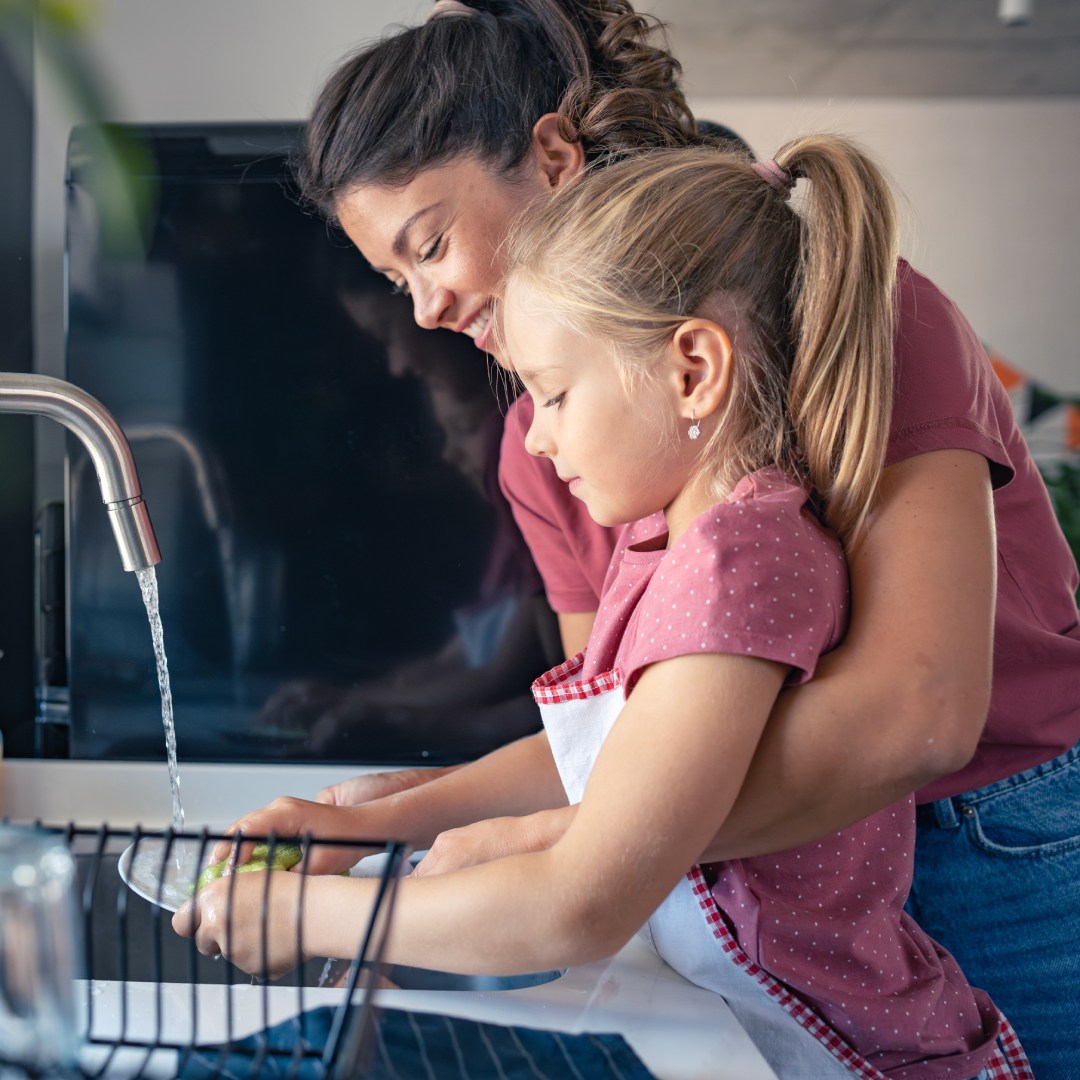 A mother helps her younger daughter at the sink while she washes off a dish under the faucet. The child wears an apron.