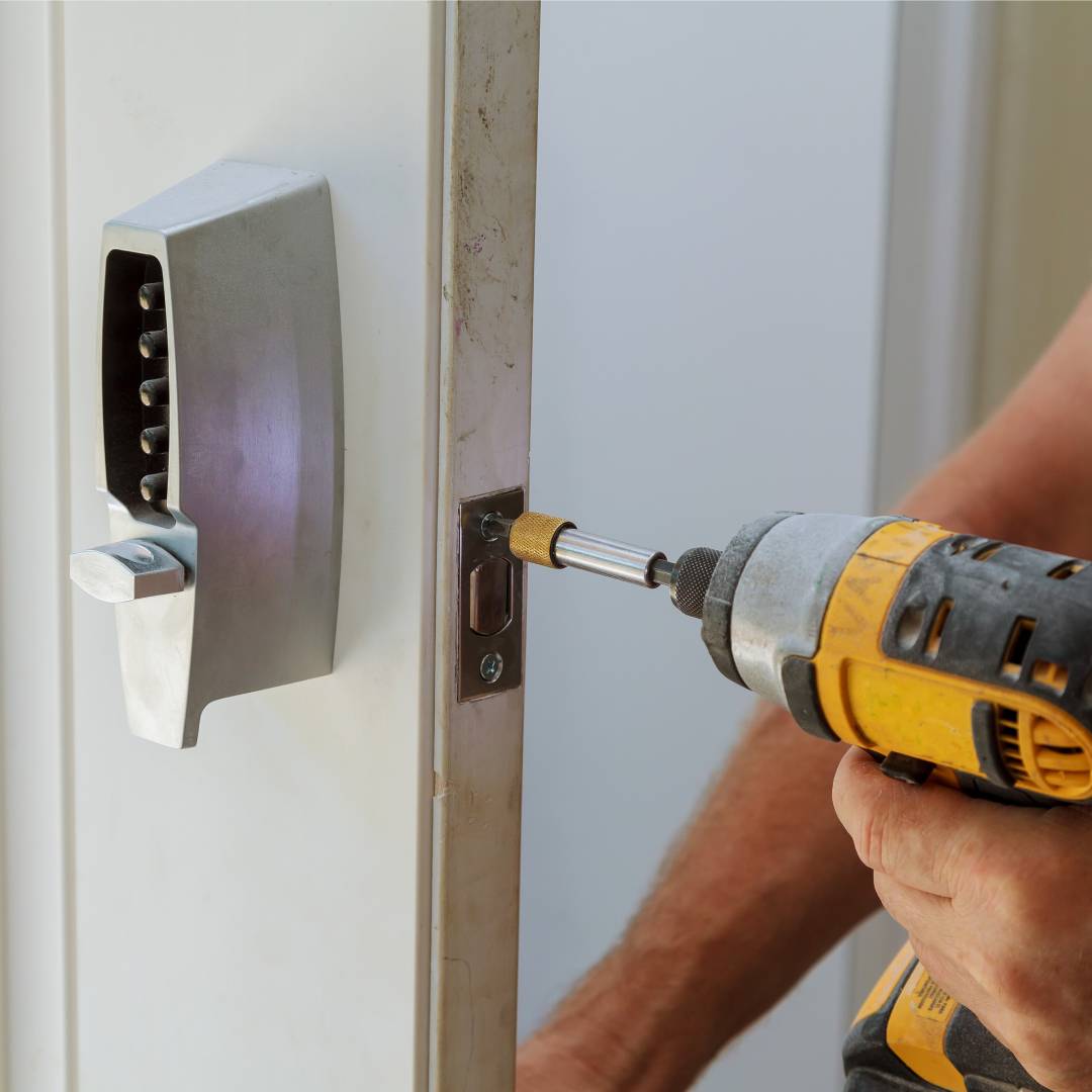 A professional locksmith working on a residential door lock with a drill. The lock has an advanced keypad.