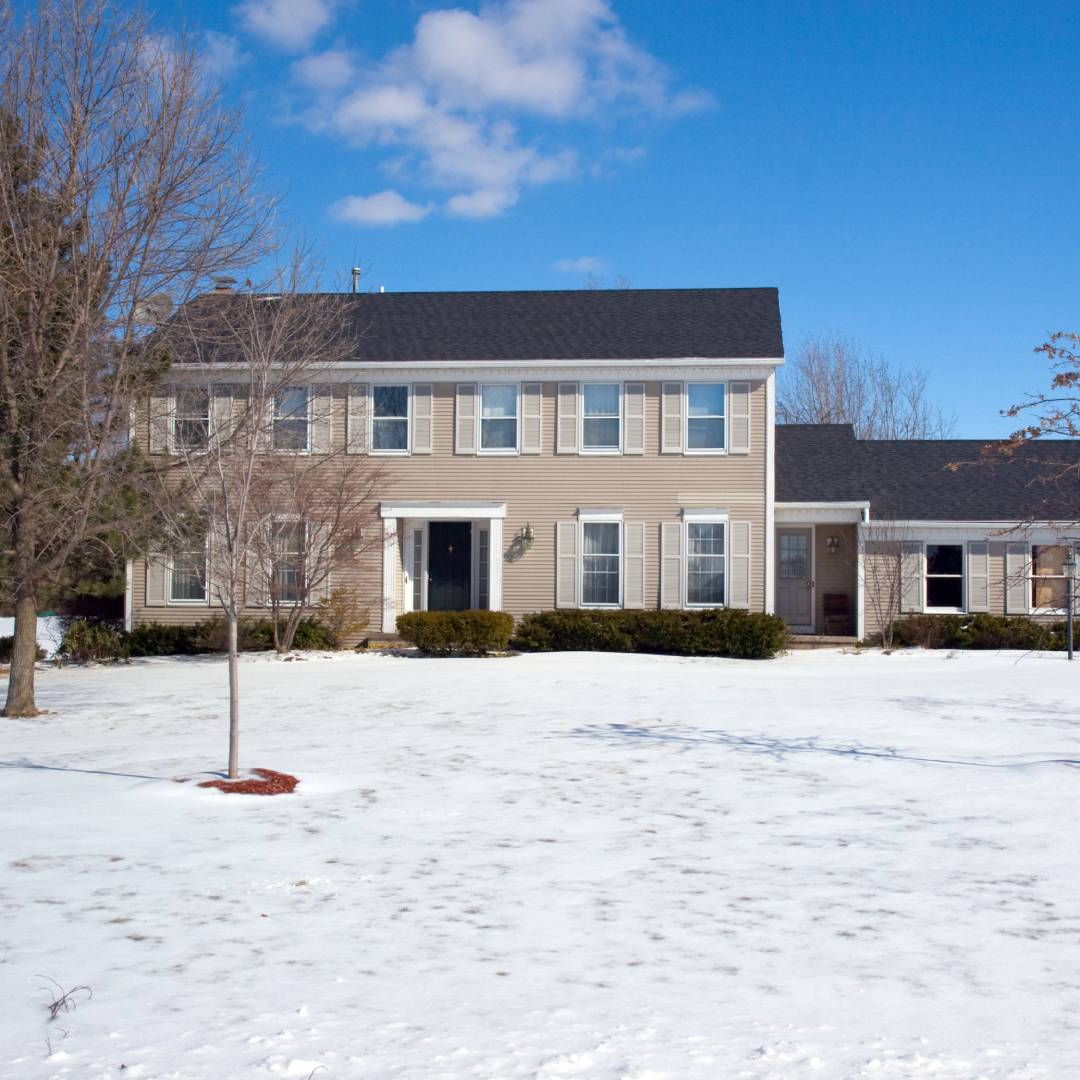 A two-story home features a vast front lawn that is covered in snow. There are several trees on the lawn.