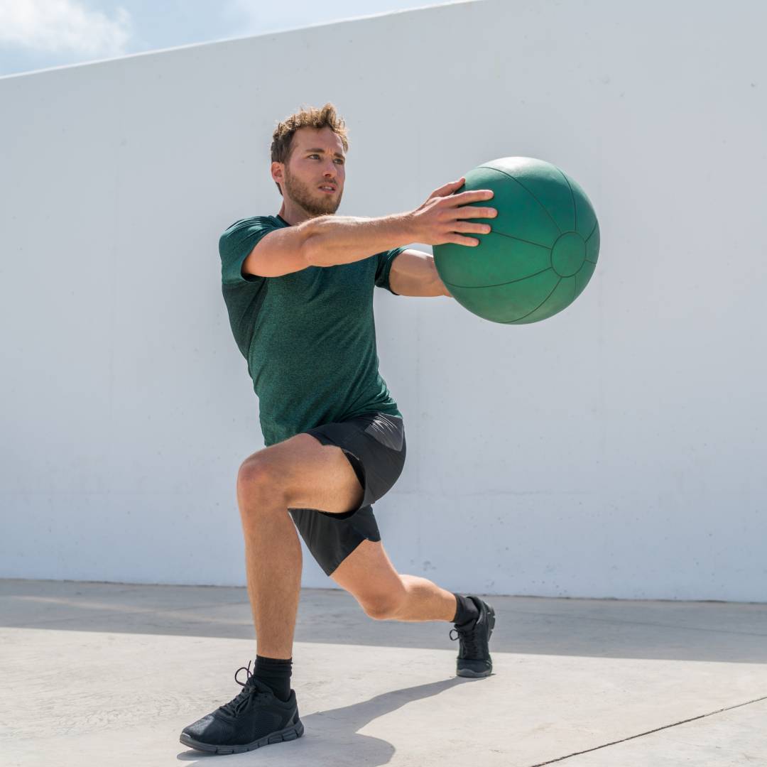 A man wearing a green T-shirt and dark gray shorts works out with a green medicine ball in front of a white wall.