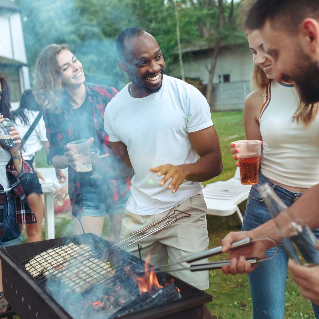 A group of young people gathered around a grill. They are holding beverages and smiling at each other.