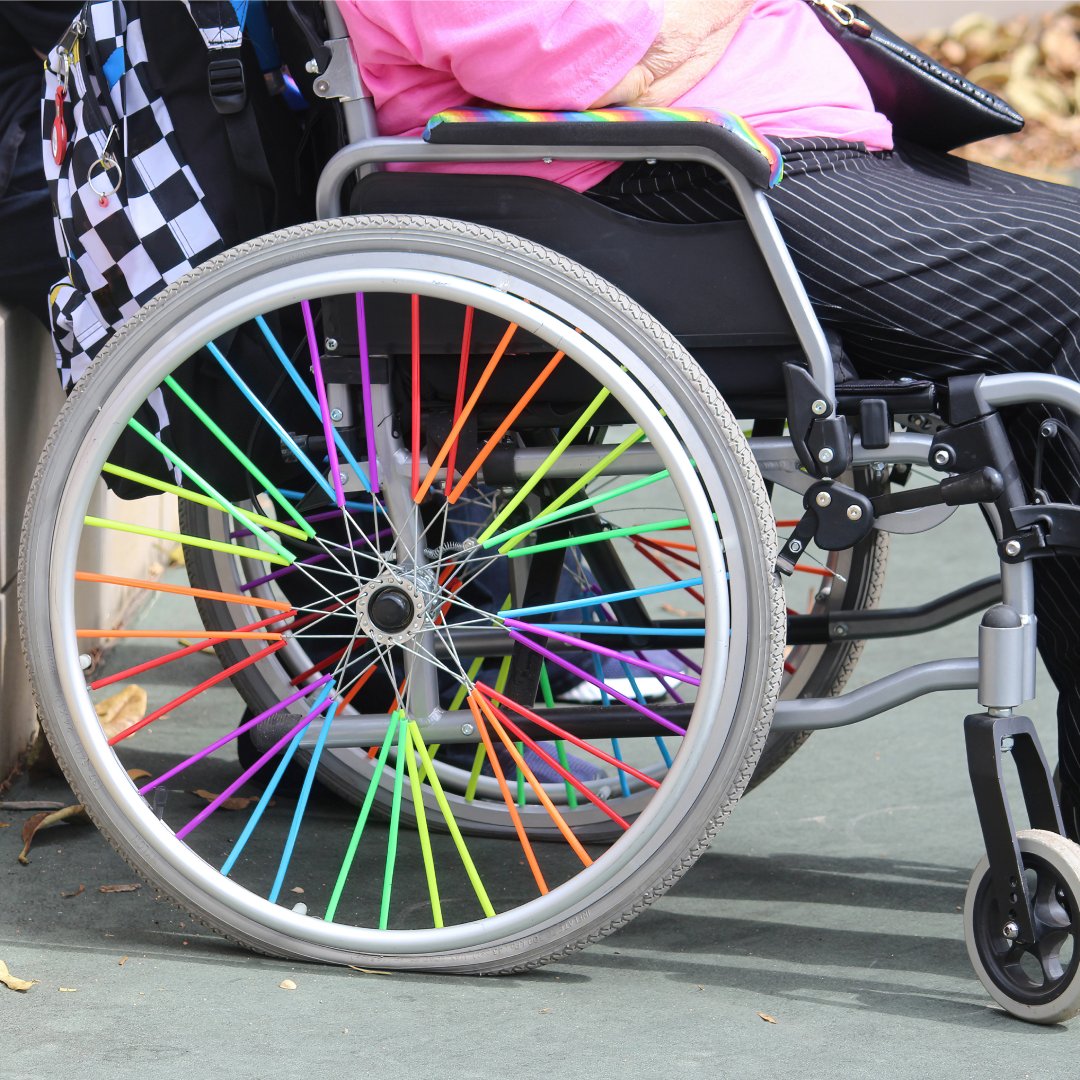 Someone sits in an eclectic wheelchair. The spokes are rainbow-colored, and there is a black and white checked backpack.