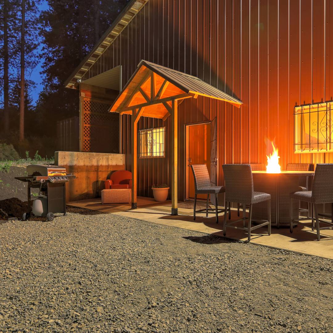 The exterior of a metal barn at night. There is a circular fire pit, outdoor furniture, and a grill in front of the barn.