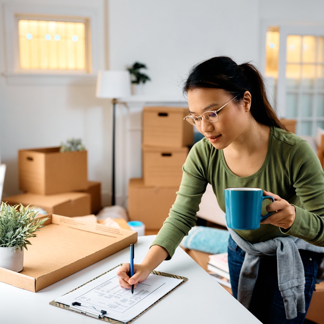 A woman in her apartment with several cardboard boxes marks a checklist on a clipboard while holding a blue mug.