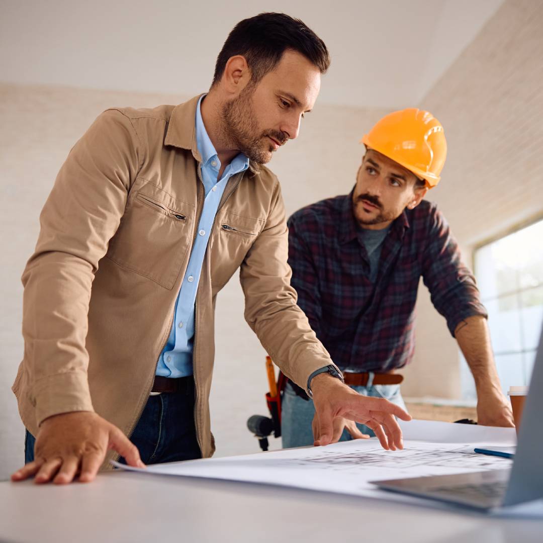 A worker wearing a yellow hard hat talks with another person while pouring over a computer and blueprints.