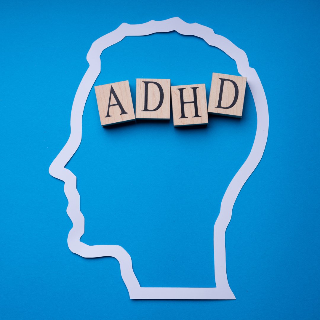 A white paper outline of a head rests on a blue background. Wooden blocks spelling "ADHD" sit inside the head.