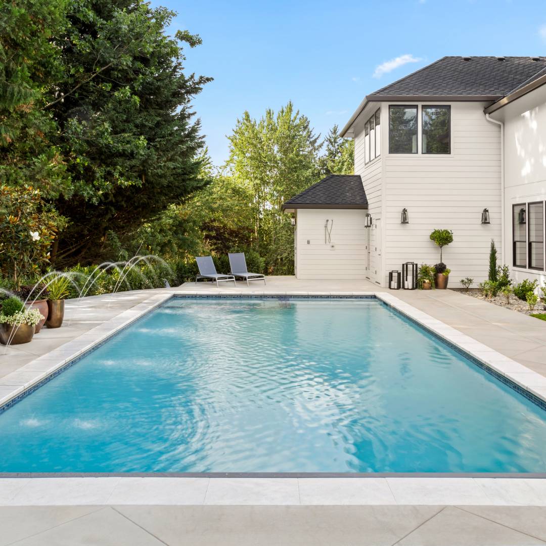 A below ground swimming pool in the backyard of a residential home with clear blue water next to a row of trees.