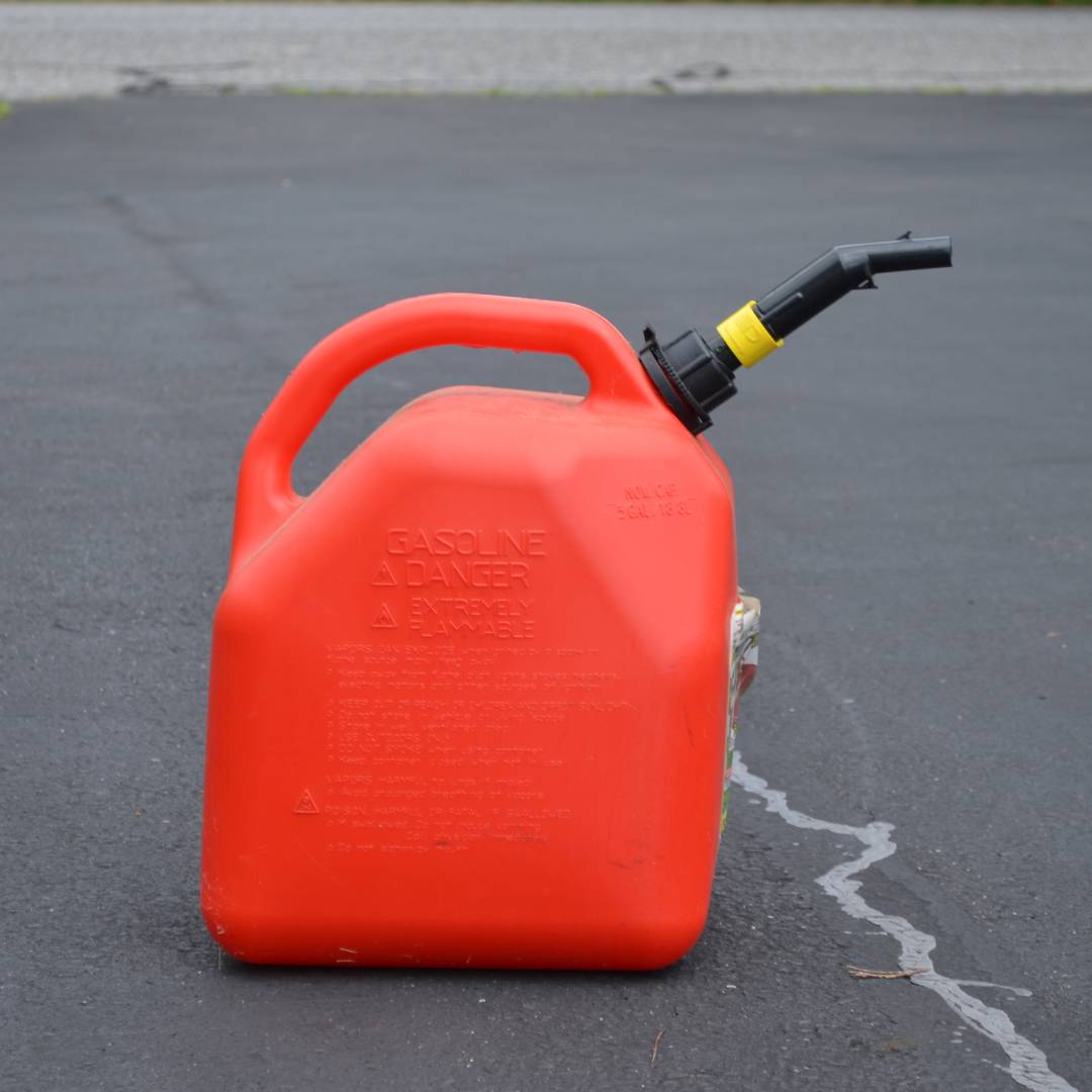 A red gasoline can with a black spout sits in a black asphalt driveway with a road in the background.