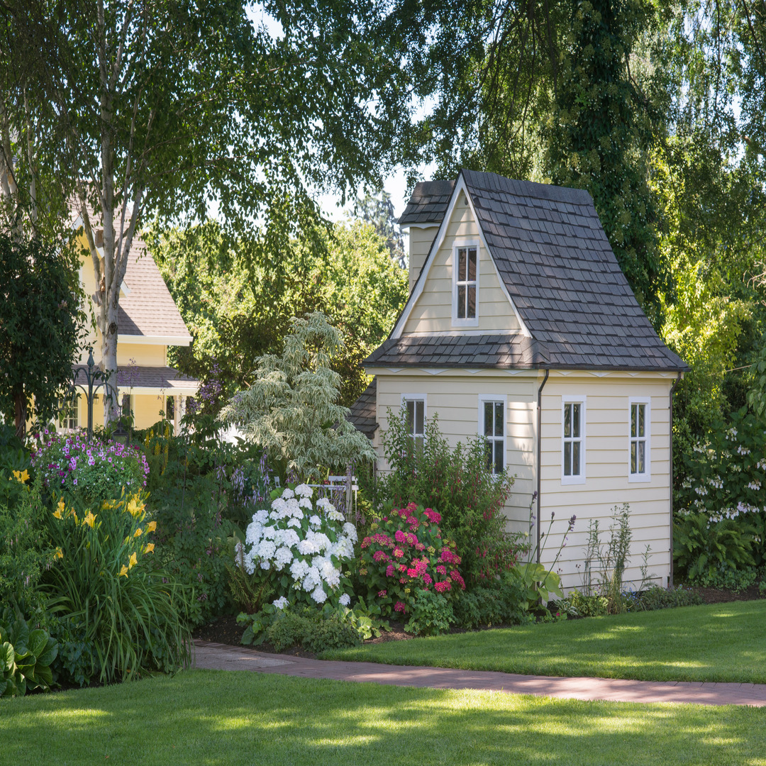 A pale yellow tiny house with a large garden filled with pink and white flowers. Another yellow house sits in the distance.