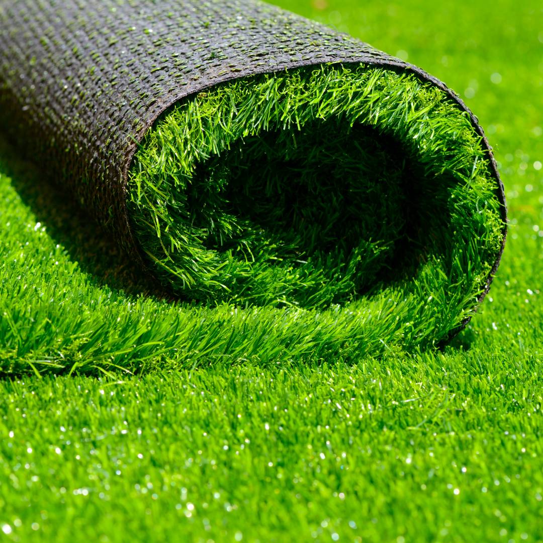 A strip of artificial turf, rolled up and being laid out on a stretch of already-placed artificial turf in the sunlight.