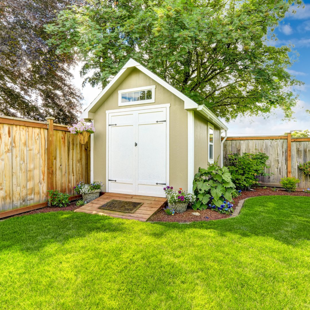 A beige shed with double doors is up against the corner of a fenced backyard with plants around the perimeter.