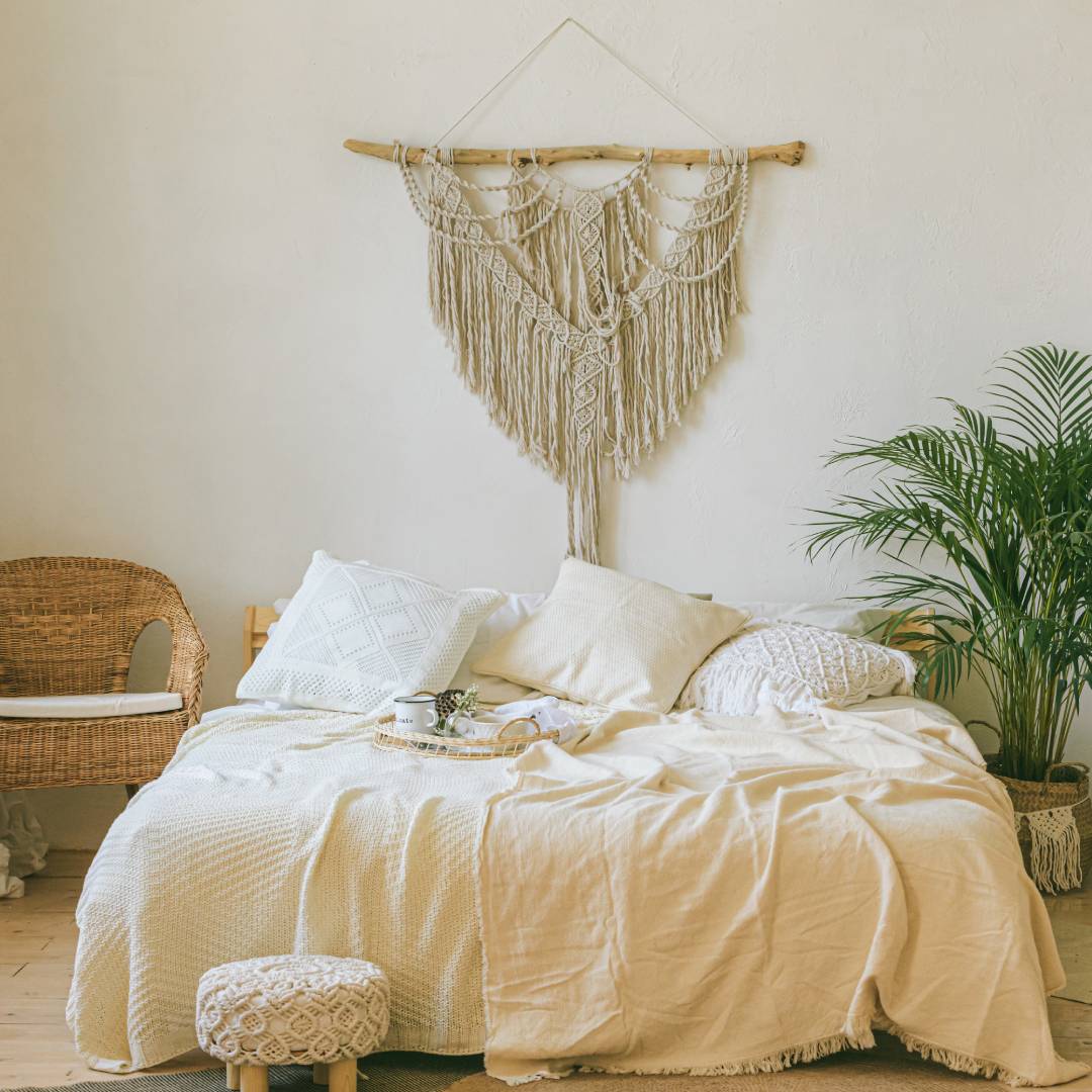 A bedroom with a large bed up against the wall. Hanging above it is a piece of woven macramé art attached to a wooden stick.