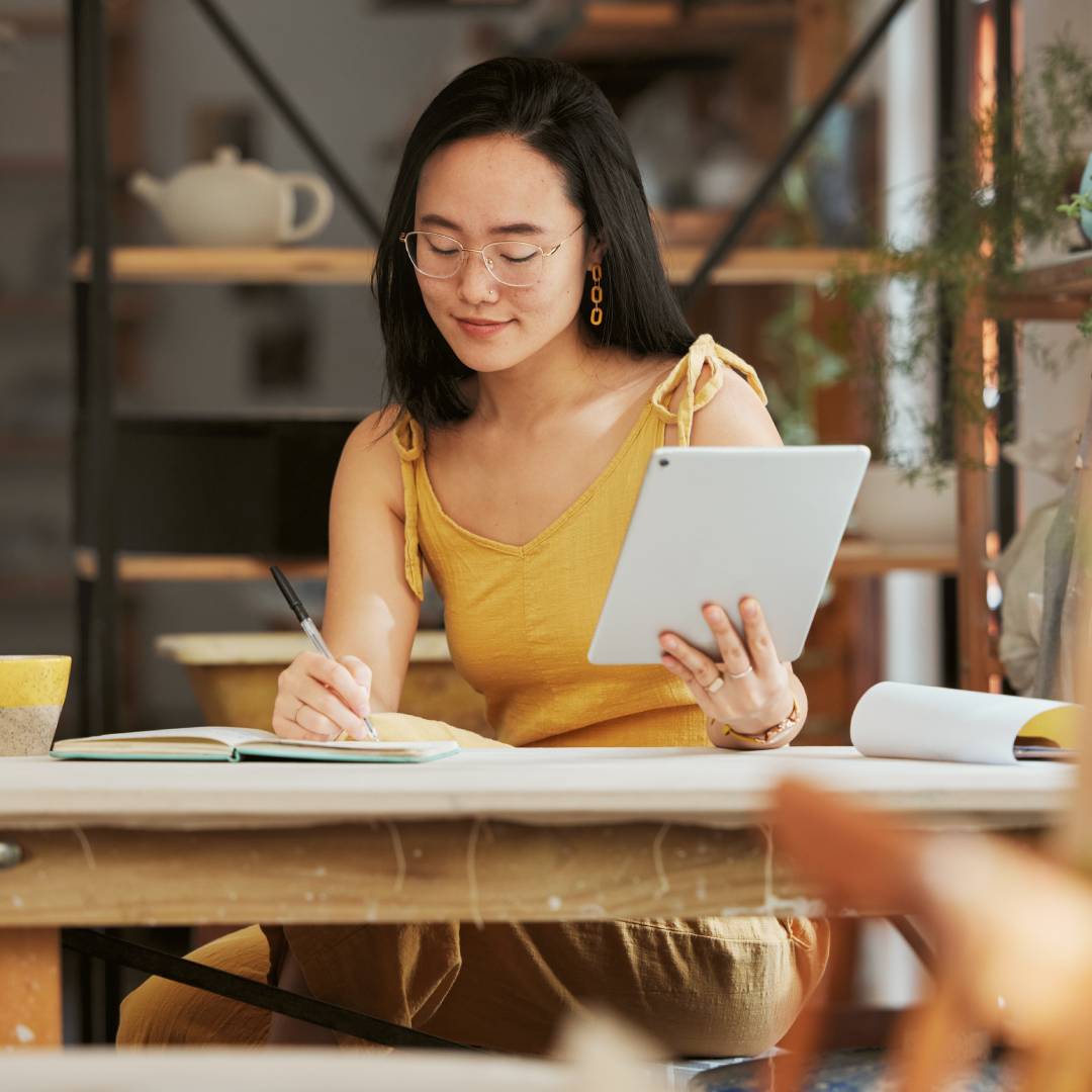 A woman sits at a desk surrounded by plants and shelves. She writes in a notebook and holds a tablet in her other hand.