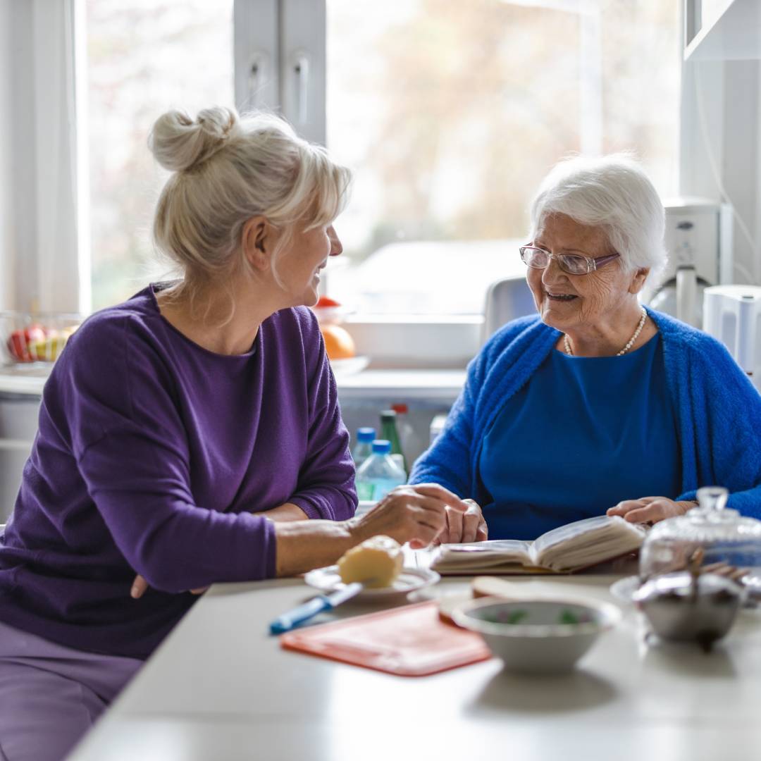 Two women, one of them older than the other, sit, smiling as they talk, at a kitchen table.