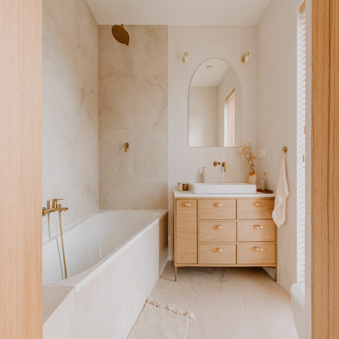 A natural-looking bathroom contains a white tub, a wooden vanity, gold fixtures, a large mirror, and marble flooring.