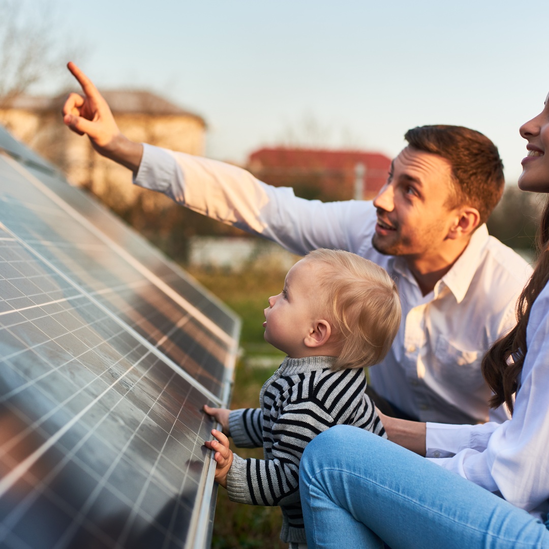 A close-up of a man and a woman in white shirts showing a blond baby a row of solar panels in a field.