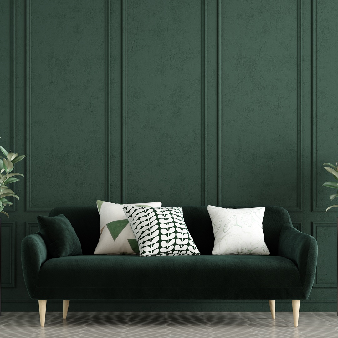 A dark green velvet couch with throw pillows on it and two plants in front of a green paneled wall.