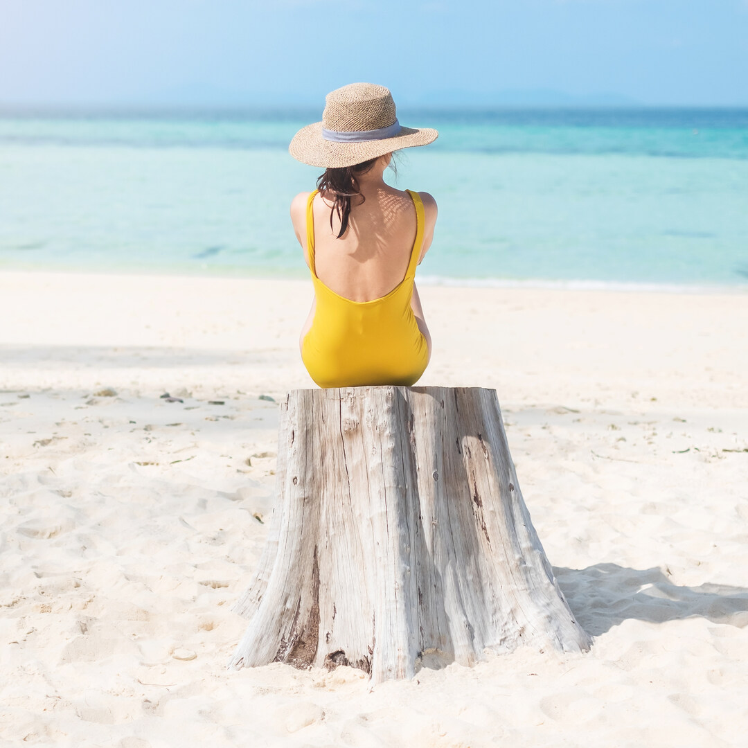 A woman in a yellow dress and a sun hat sitting on a stump on a beach looking at the bright-blue water of the ocean.