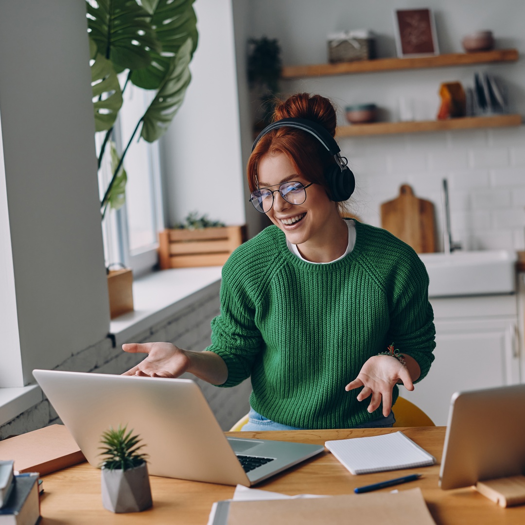 A young woman wearing glasses and a green sweater smiles while sitting at a desk with a silver laptop and tablet.