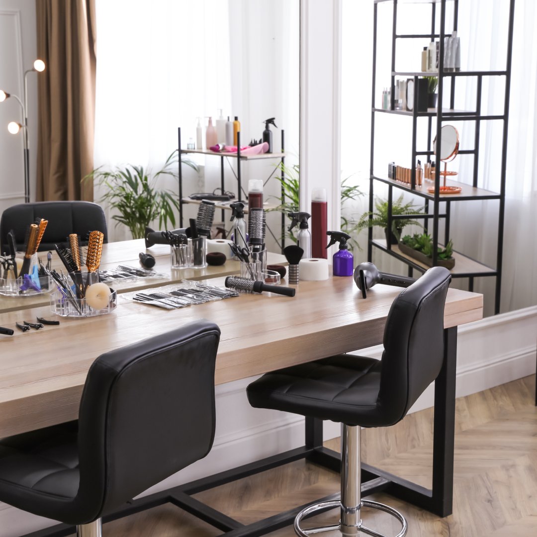 A full service beauty salon with two black service chairs situated at a work desk. There are indoor plants near the window.
