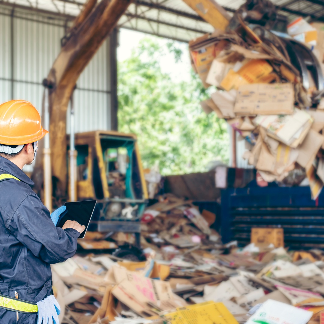 A recycling industry professional in a yellow hard hat watching an excavator dump cardboard waste into a warehouse.