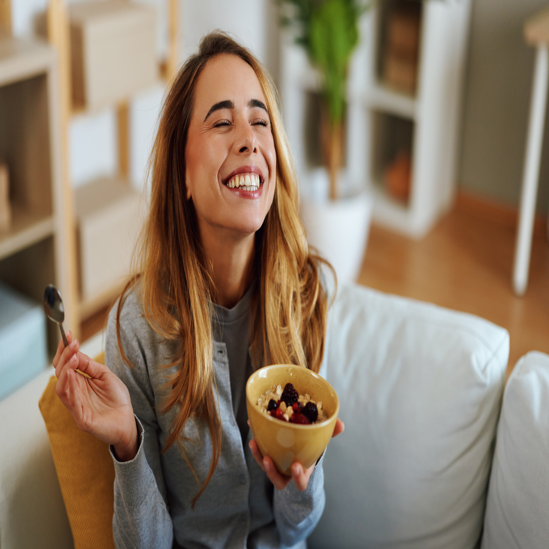 A young woman is smiling while sitting on the couch. She's holding a bowl in one hand and a spoon in the other.