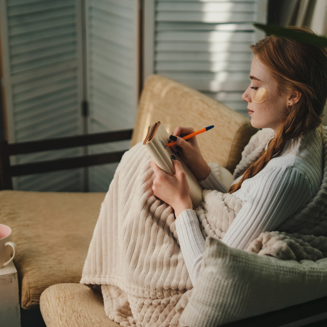 A woman sits on a loveseat with her knees to her chest under a blanket, writing in a journal in front of her laptop and a mug.