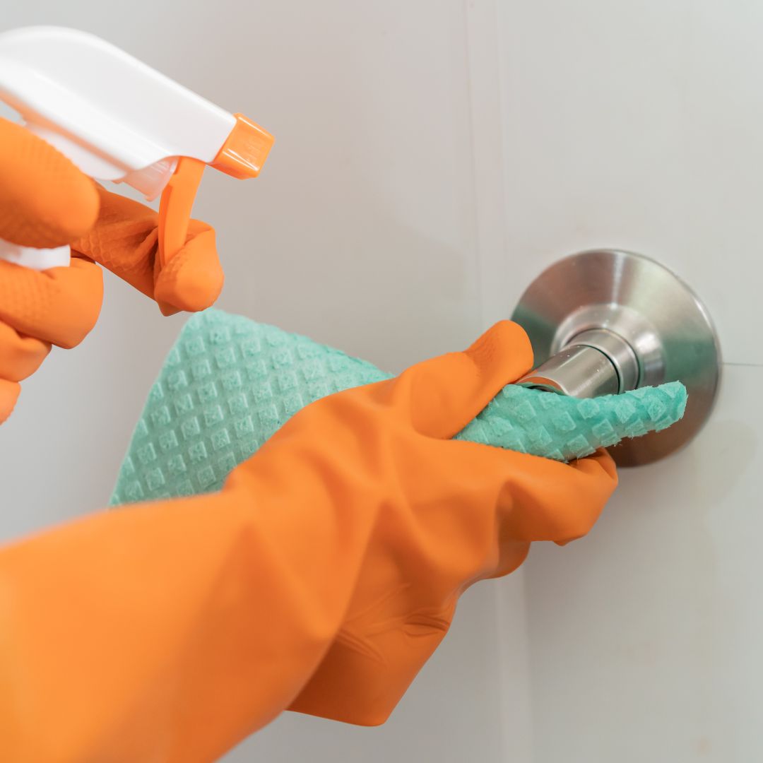 Close-up of a person's hands spraying and wiping off a doorknob while wearing bright orange gloves.