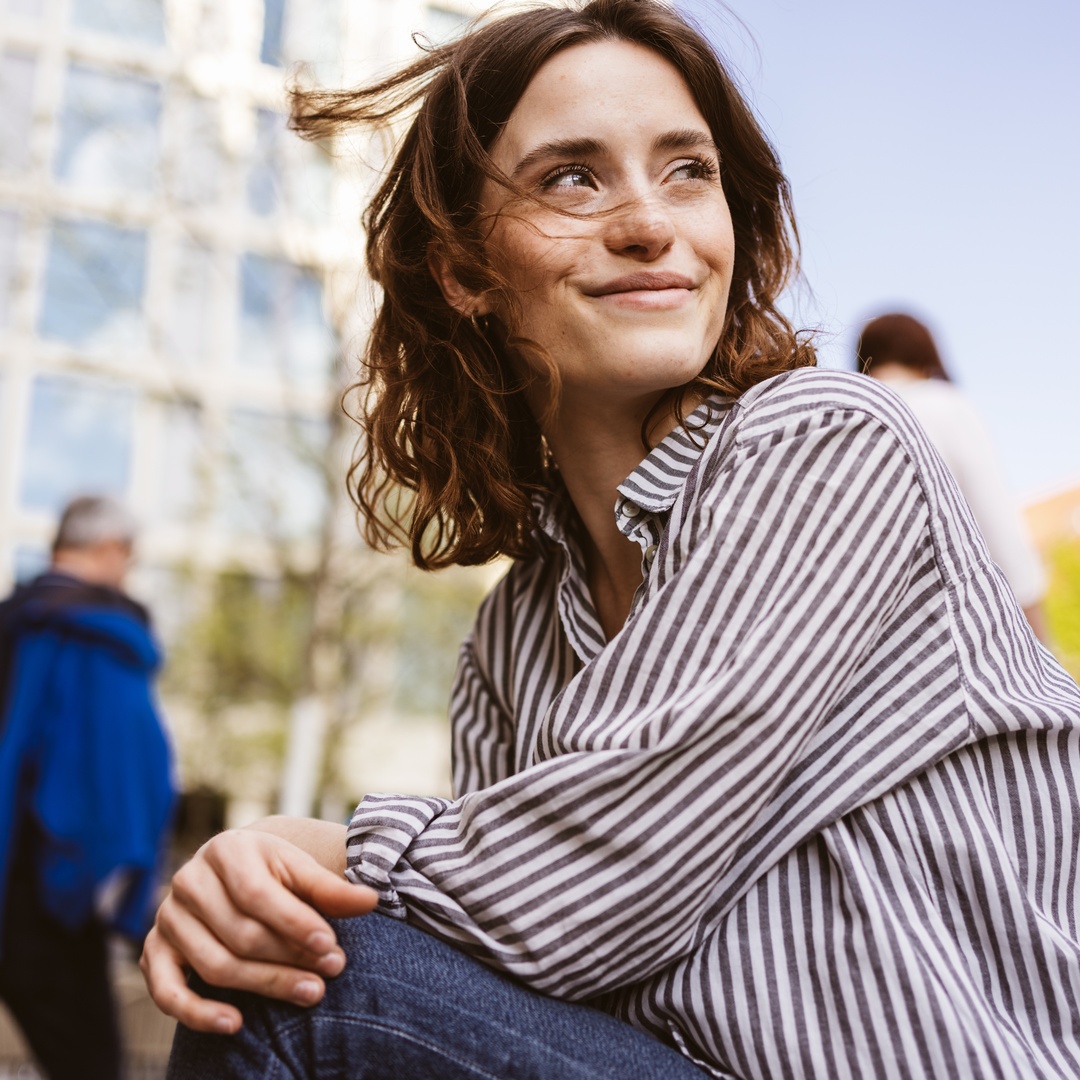 A smiling brunette woman in a black-and-white striped shirt sitting on a shaded step in an outdoor courtyard.