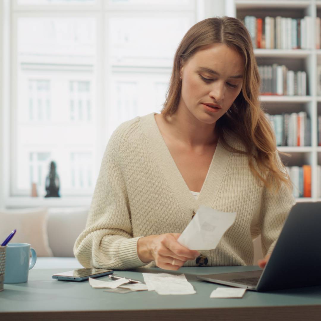A young blonde woman sitting at a desk in front of a laptop and looking down at one of several paper receipts.