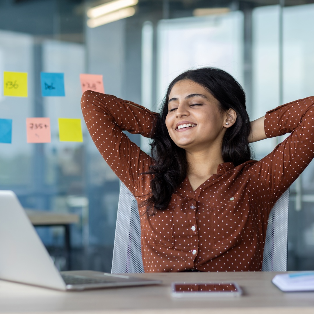 A woman sits back and relaxes with her hands behind her head in her office. She has a laptop in front of her at her desk.