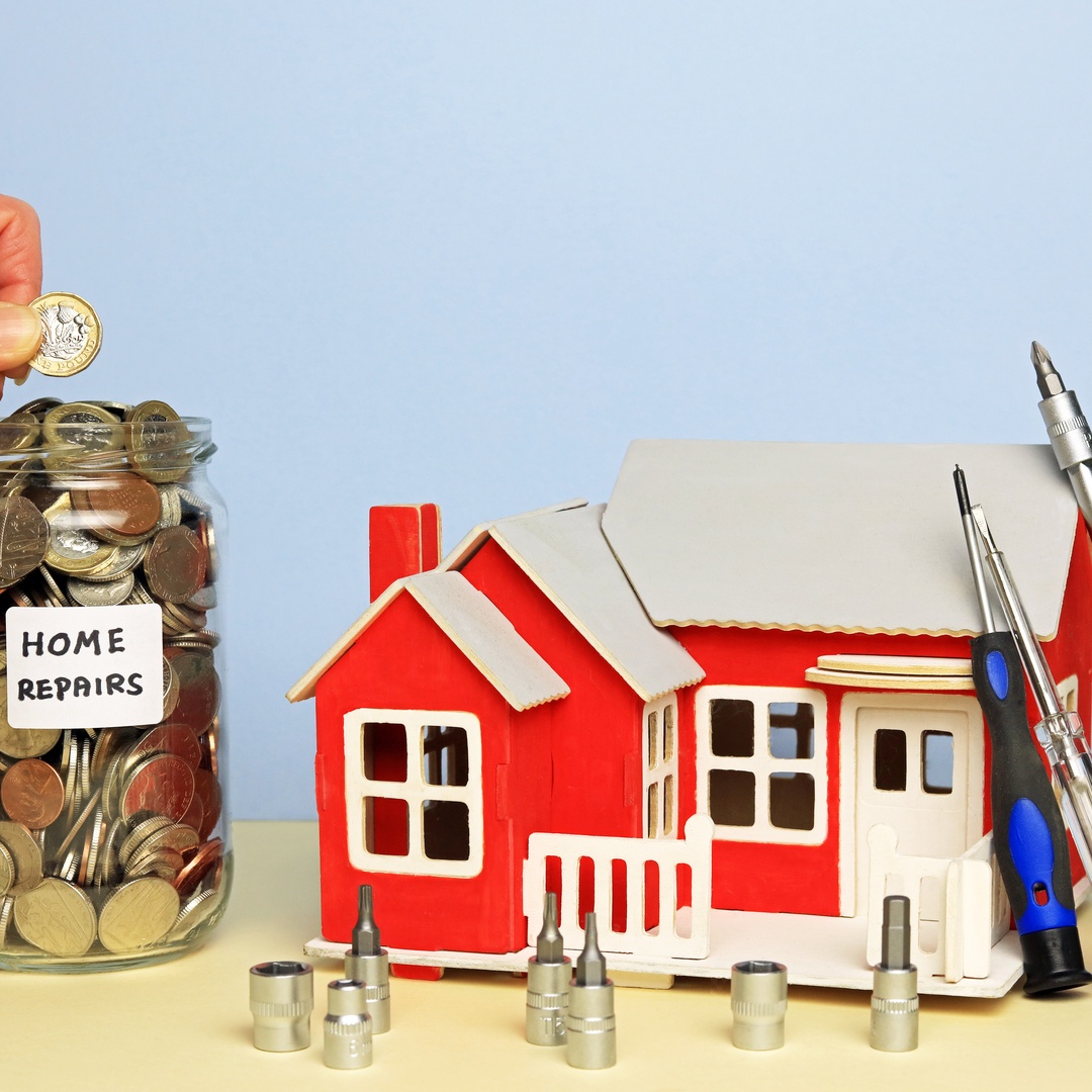 A person's hand puts coins in a jar labeled "Home Repairs." A small model home sits next to the jar with tools leaning against it.